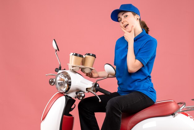 Vista frontal de mensajería femenina sentada en bicicleta con tazas de café en el trabajo de color rosa uniforme de entrega de alimentos trabajador