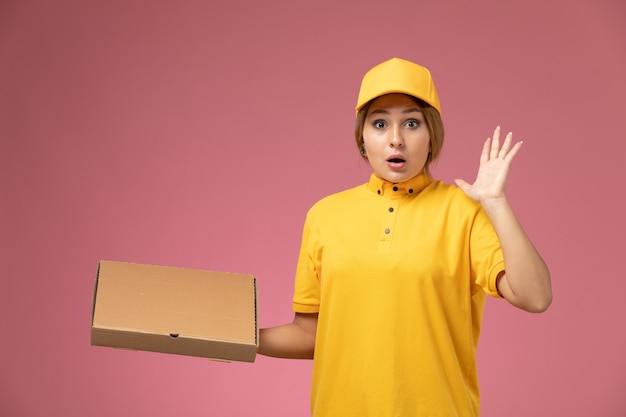Vista frontal de mensajería femenina en uniforme amarillo capa amarilla sosteniendo la caja de entrega de alimentos en el escritorio rosa entrega uniforme color femenino