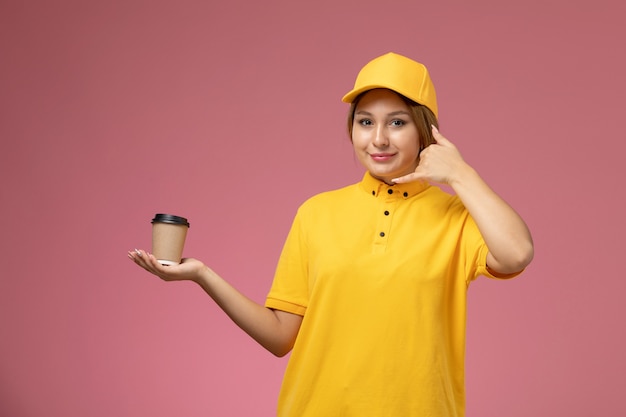 Vista frontal de mensajería femenina en uniforme amarillo capa amarilla sosteniendo una taza de café marrón de plástico en el escritorio rosa entrega uniforme color de niña femenina