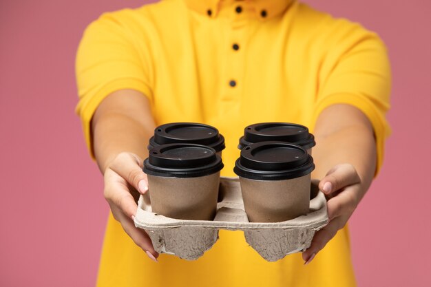 Vista frontal de mensajería femenina en uniforme amarillo capa amarilla sosteniendo tazas de café de plástico en el escritorio rosa trabajo de entrega uniforme trabajo de color