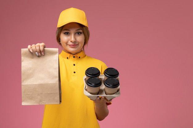 Vista frontal de mensajería femenina en uniforme amarillo capa amarilla sosteniendo tazas de café de plástico paquete de alimentos sobre fondo rosa trabajo de entrega uniforme trabajo de color