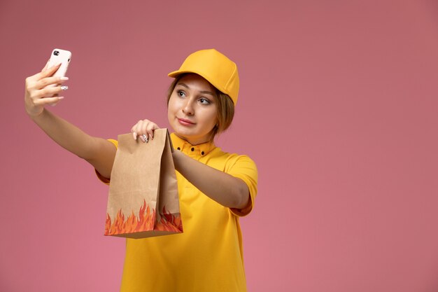 Vista frontal de mensajería femenina en uniforme amarillo capa amarilla takign un selfie con paquete de comida sobre fondo rosa trabajo de entrega uniforme trabajo de color