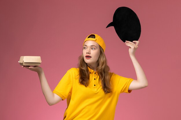 Vista frontal de mensajería femenina en uniforme amarillo y capa con cartel negro con tazón en la pared rosa servicio de entrega uniforme empresa gitl trabajador