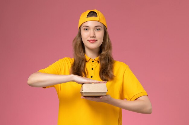 Vista frontal de mensajería femenina en uniforme amarillo y capa con paquete de comida de entrega en la pared rosa servicio de entrega uniforme empresa trabajo trabajador