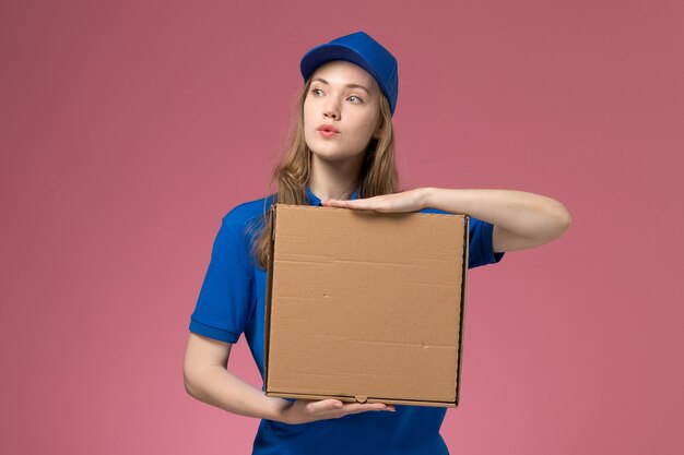 Vista frontal de mensajería femenina en uniforme azul con caja de entrega de alimentos en la empresa de uniforme de servicio de fondo rosa