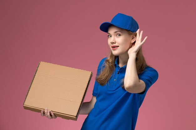 Vista frontal de mensajería femenina en uniforme azul sosteniendo la caja de comida tratando de escuchar sobre fondo rosa empresa uniforme de servicio de trabajo