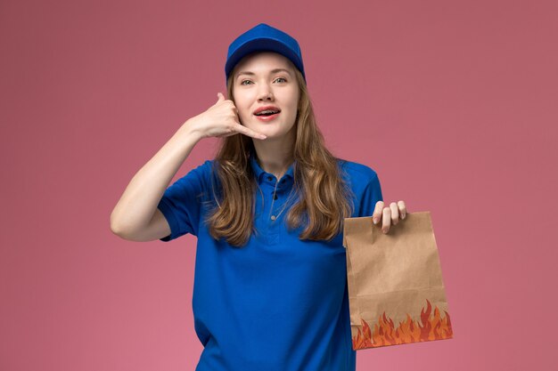 Vista frontal de mensajería femenina en uniforme azul sosteniendo el paquete de alimentos que muestra el gesto de llamada telefónica en la empresa de uniforme de servicio de trabajador de trabajo de escritorio rosa