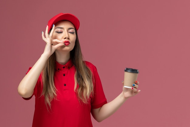 Vista frontal de mensajería femenina en uniforme rojo y capa sosteniendo la taza de café de entrega sobre fondo rosa servicio de entrega uniforme trabajo niña trabajadora