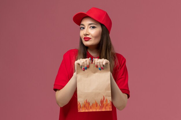 Vista frontal de mensajería femenina en uniforme rojo con paquete de comida de papel sobre fondo rosa trabajo de empresa uniforme de entrega de servicio