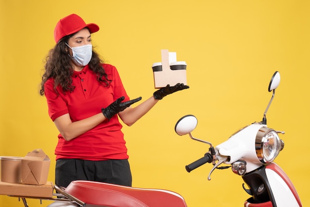 Vista frontal de mensajería femenina en uniforme rojo sosteniendo café sobre fondo amarillo trabajador entrega covid- pandemia servicio uniforme de trabajo