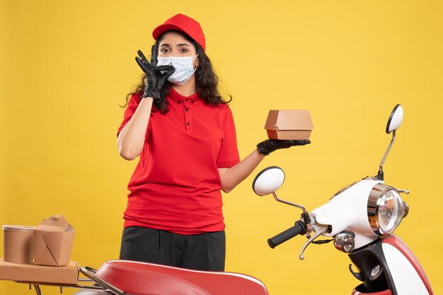 Vista frontal de mensajería femenina en uniforme rojo sosteniendo un pequeño paquete de comida sobre fondo amarillo entrega covid- pandemia de trabajador uniforme de trabajo