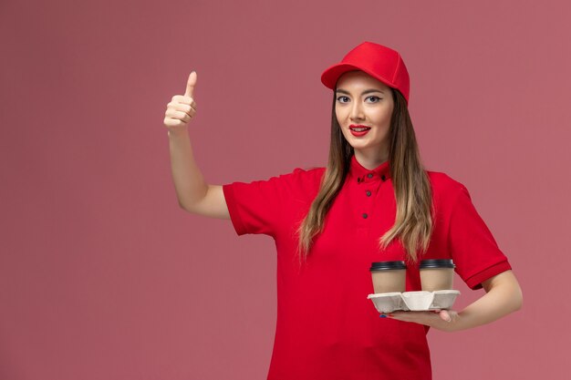 Vista frontal de mensajería femenina en uniforme rojo sosteniendo tazas de café de entrega marrón sonriendo sobre fondo rosa uniforme de entrega de trabajo de servicio