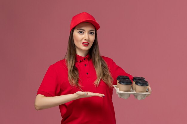 Vista frontal de mensajería femenina en uniforme rojo sosteniendo tazas de café de entrega sobre fondo rosa trabajador de trabajo uniforme de entrega de servicio