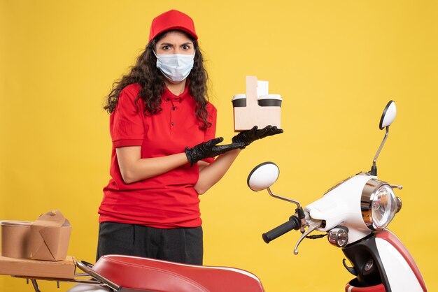 Vista frontal de mensajería femenina en uniforme rojo sosteniendo tazas de café sobre fondo amarillo entrega de trabajadores servicio uniforme de trabajo pandemia covid