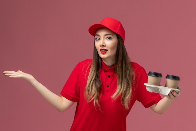 Vista frontal de mensajería femenina en uniforme rojo con tazas de café de entrega marrón sobre fondo rosa servicio de entrega uniforme trabajador trabajo empresa femenina