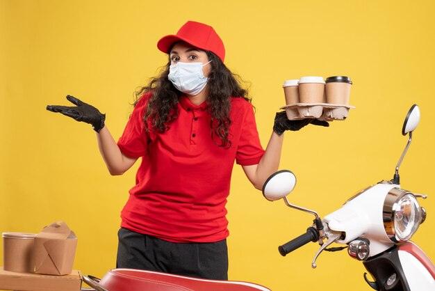 Vista frontal de mensajería femenina en uniforme rojo con tazas de café sobre un fondo amarillo trabajador entrega covid- trabajo de virus de servicio pandémico