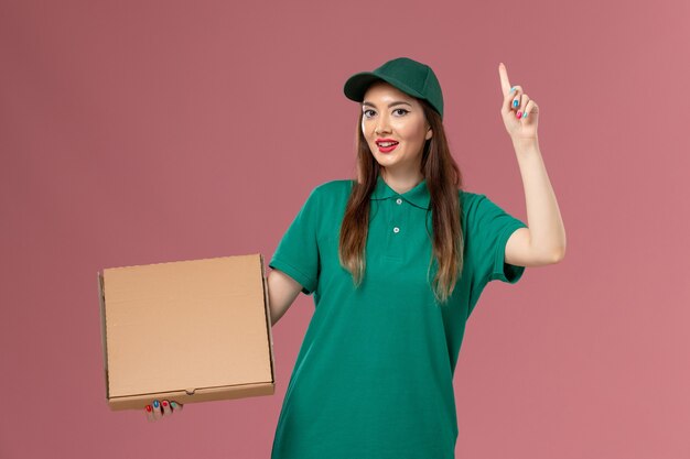 Vista frontal de mensajería femenina en uniforme verde con caja de comida en la empresa de entrega uniforme de servicio de trabajo de pared rosa