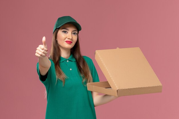 Vista frontal de mensajería femenina en uniforme verde con caja de entrega de alimentos en la pared rosa servicio de la empresa trabajo de entrega uniforme