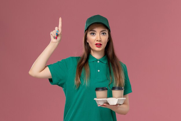 Vista frontal de mensajería femenina en uniforme verde y capa sosteniendo tazas de café de entrega en la pared rosa empresa servicio trabajo uniforme entrega trabajadora mujer