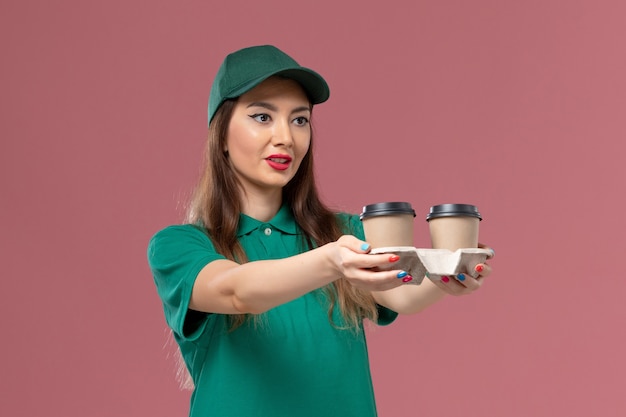 Vista frontal de mensajería femenina en uniforme verde y capa sosteniendo tazas de café de entrega en la pared rosa servicio de la empresa trabajo uniforme entrega femenina