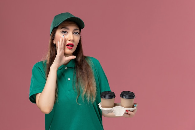 Vista frontal de mensajería femenina en uniforme verde y capa sosteniendo tazas de café de entrega susurrando en la pared rosa empresa servicio trabajo uniforme entrega trabajadora mujer trabajo niña