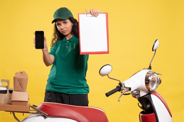 Vista frontal de mensajería femenina en uniforme verde con teléfono y nota de archivo sobre el fondo amarillo trabajador de servicio trabajo entrega trabajo comida