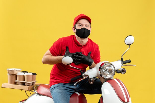 Vista frontal del mensajero agradecido hombre vestido con blusa roja y guantes de sombrero en máscara médica entregando orden sentado en scooter