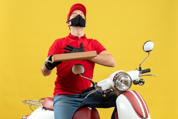 Vista frontal del mensajero agradecido hombre vestido con blusa roja y guantes de sombrero en máscara médica sentado en scooter mostrando orden