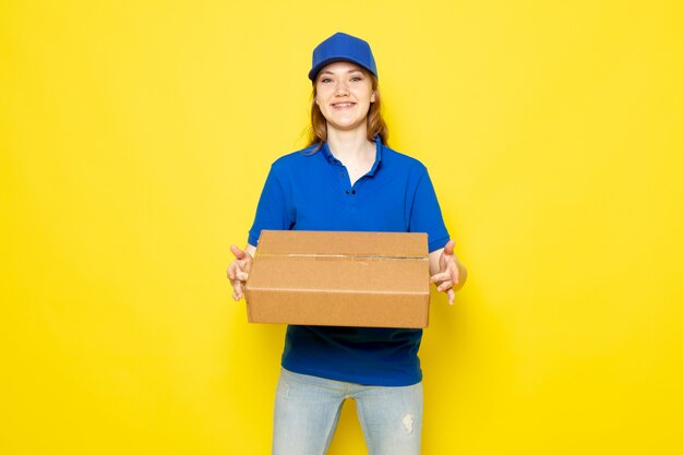Una vista frontal mensajero atractivo femenino en polo azul gorra azul y jeans sonriendo sosteniendo el paquete en el trabajo de servicio de alimentos de fondo amarillo