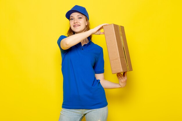 Una vista frontal mensajero atractivo femenino en polo azul gorra azul y jeans sosteniendo paquete sonriendo en el trabajo de servicio de alimentos de fondo amarillo