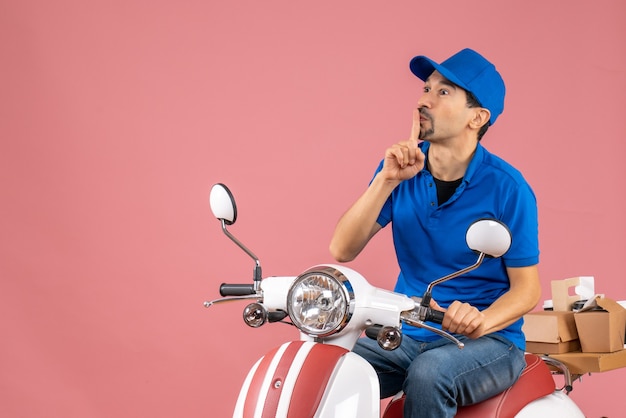 Vista frontal del mensajero chico con sombrero sentado en scooter haciendo gesto de silencio sobre fondo melocotón pastel