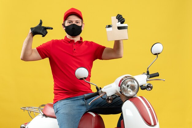 Vista frontal del mensajero confiado hombre vestido con blusa roja y guantes de sombrero en máscara médica sentado en scooter mostrando pedidos