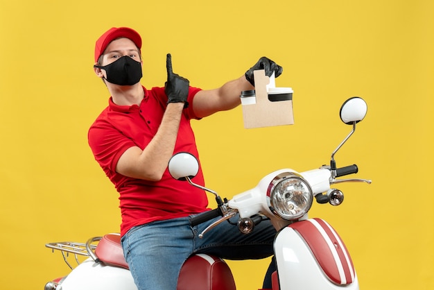 Vista frontal del mensajero confiado hombre vestido con blusa roja y guantes de sombrero en máscara médica sentado en scooter sosteniendo órdenes mostrando uno