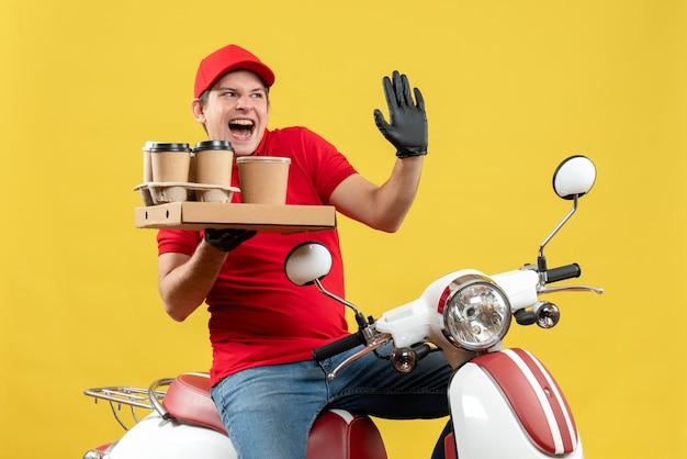 Vista frontal del mensajero emocional hombre vestido con blusa roja y guantes de sombrero en máscara médica entregando orden sentado en scooter con órdenes