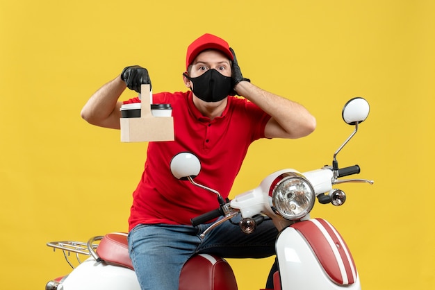 Vista frontal del mensajero enfocado hombre vestido con blusa roja y guantes de sombrero en máscara médica sentado en scooter mostrando pedidos