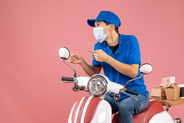 Vista frontal del mensajero enfocado en máscara médica con sombrero sentado en scooter sobre fondo de melocotón pastel