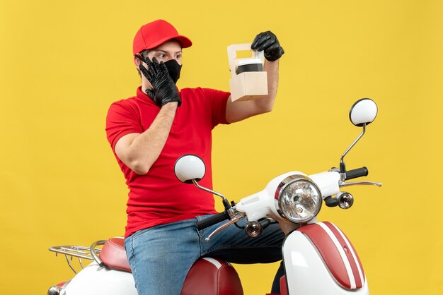 Foto gratuita vista frontal del mensajero enfocado sorprendido hombre vestido con blusa roja y guantes de sombrero en máscara médica sentado en scooter