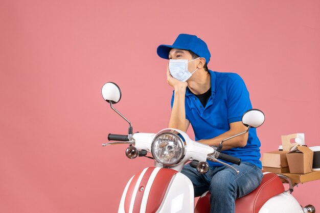 Vista frontal del mensajero de ensueño en máscara médica con sombrero sentado en scooter sobre fondo de melocotón pastel