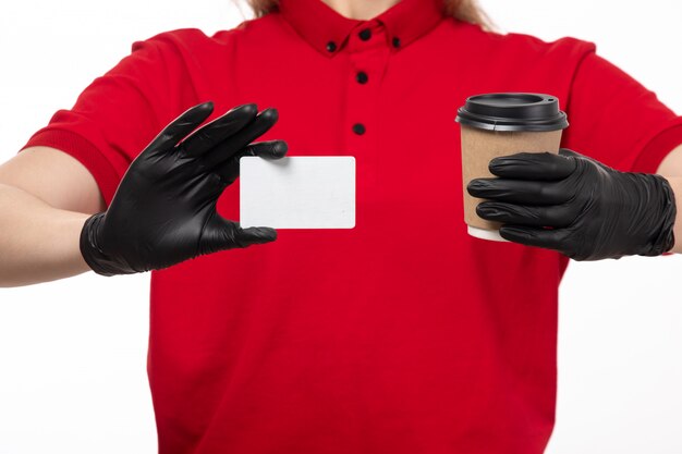 Una vista frontal mensajero femenino en camisa roja guantes negros sosteniendo la taza con café y tarjeta blanca sobre blanco
