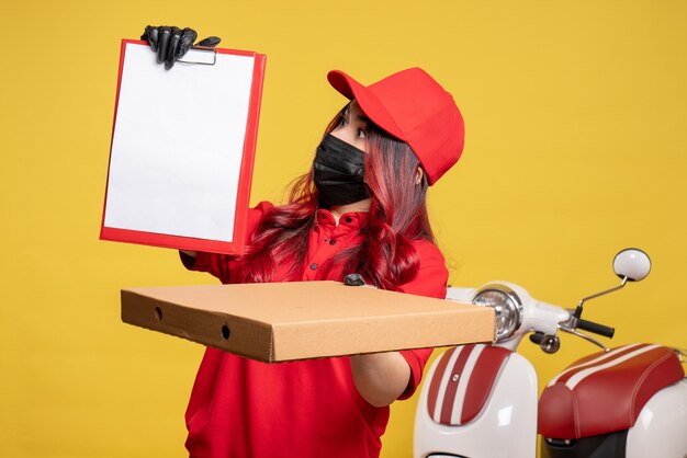 Vista frontal del mensajero femenino en máscara con caja de comida de entrega y nota de archivo en la pared amarilla