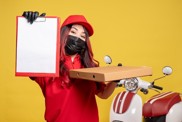 Foto gratuita vista frontal del mensajero femenino en máscara con caja de comida de entrega y nota de archivo en la pared amarilla