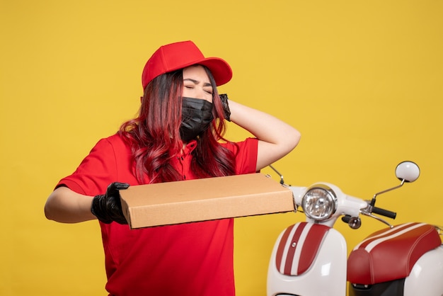 Vista frontal del mensajero femenino en máscara con caja de comida de entrega en la pared amarilla