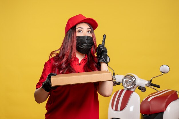 Vista frontal del mensajero femenino en máscara con caja de comida de entrega en la pared amarilla