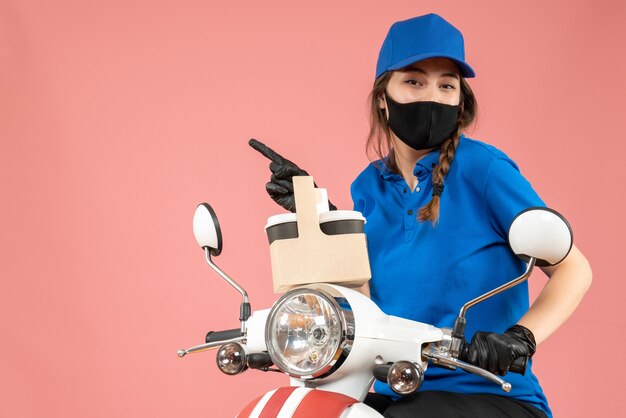 Vista frontal del mensajero femenino sonriente con máscara médica negra y guantes entregando pedidos sobre fondo melocotón