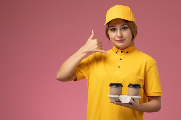 Vista frontal mensajero femenino en uniforme amarillo capa amarilla sosteniendo una taza de café marrón de plástico en el escritorio rosa uniforme femenino de entrega