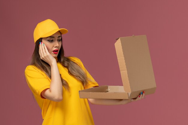 Vista frontal del mensajero femenino en uniforme amarillo y gorra con caja de comida abierta en la pared rosa claro