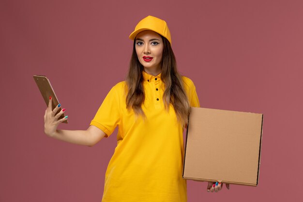 Vista frontal del mensajero femenino en uniforme amarillo y gorra con caja de comida y bloc de notas en la pared rosa