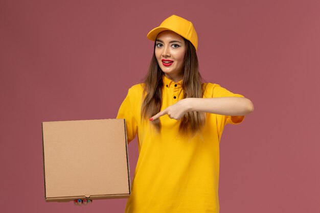 Vista frontal del mensajero femenino en uniforme amarillo y gorra con caja de comida en la pared rosa claro