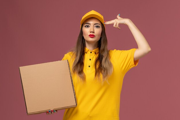 Vista frontal del mensajero femenino en uniforme amarillo y gorra con caja de comida en la pared rosa claro