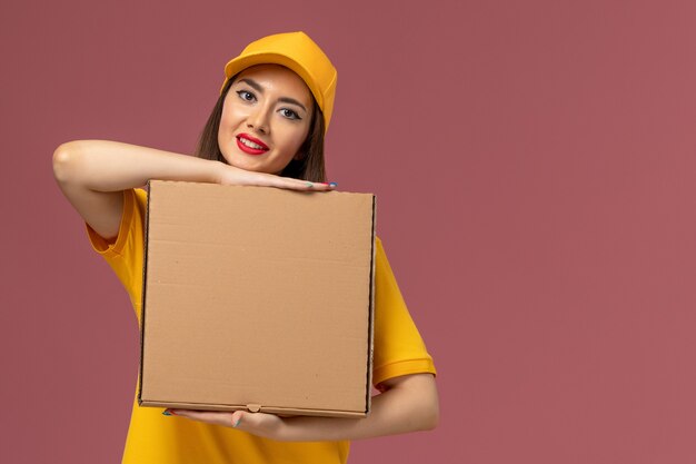 Vista frontal del mensajero femenino en uniforme amarillo y gorra con caja de comida en la pared rosa claro